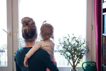 mother and daughter look through the window, toddler girl on the hands of woman, authentic home interior © skypictom