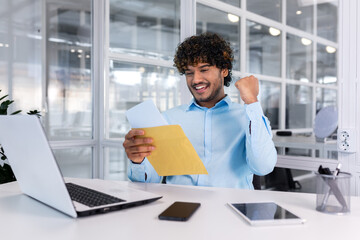 Businessman celebrating victory and success, receiving mail envelope with good news notification, hispanic reading document and happy holding hand up triumph gesture, man at workplace inside office.