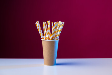 Disposable cardboard biodegradable cup on a white table with a straw against a red background.