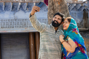 Fototapeta na wymiar Young happy rural indian couple wearing traditional outfit sitting on bed taking selfie picture with smartphone .
