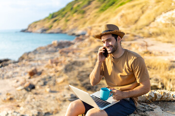 Caucasian man freelancer sitting on coastline hill working on laptop computer and talking mobile phone. Handsome guy hiking on mountain cliff on summer vacation. Digital nomad and solo travel concept.