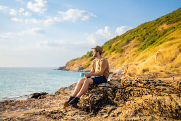 Caucasian man working outdoor on laptop computer and drinking coffee during sitting on coastline hill. Handsome guy hiking on mountain cliff on summer vacation. Digital nomad and solo travel concept.