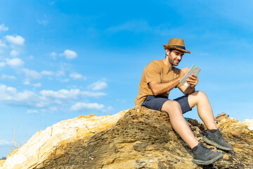 Caucasian man traveler writing journey memories in diary book on coastline hill at sunset. Handsome guy enjoy outdoor lifestyle hiking on island mountain on summer vacation. Solo travel concept.