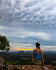 A girl sits on top of Mount Ngungun and admires a rare phenomenon, the spectacular Mammatus clouds formed during sunset in Glass House Mountains National Park, Queensland, Australia