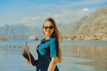 Fototapeta premium A woman in sun glasses and a hat stands against the backdrop of the sea and mountains. Vacation and outdoor recreation. Girl with long hair on vacation