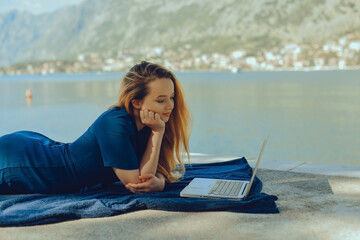 Girl freelancer lies on the background of the mountains with a laptop, remote work. A woman looks at the screen and writes something on the beach in the summer.