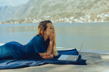 Girl freelancer lies on the background of the mountains with a laptop, remote work. A woman looks at the screen and writes something on the beach in the summer.