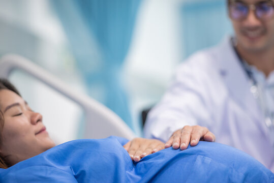 Man Male Obstetrician Doctor In White Uniform Coat Gown With Stethoscope To Check Womb Stomach In Delivery Room. Gynecologist In Gown Coat Sit And Hand Touch Check Womb Pregnant Abdomen Mother.