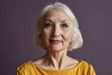Minimal close up portrait of of elegant senior woman wearing makeup and looking at camera against dusty purple background