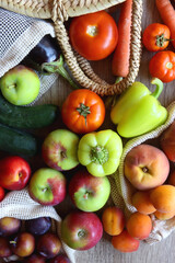 Straw bag and reusable fabric bags filled with various healthy fruit and vegetables. Wooden background, top view.