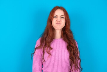 young woman wearing pink sweater over blue background puffing cheeks with funny face. Mouth inflated with air, crazy expression. - obrazy, fototapety, plakaty