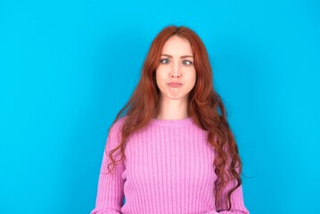 Fototapeta premium young woman wearing pink sweater over blue background crosses eyes, puts lips, makes grimace with awkward expression has fun alone, plays fool.