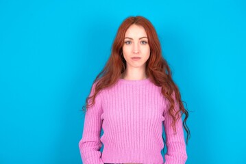 Joyful young woman wearing pink sweater over blue background looking to the camera, thinking about something. Both arms down, neutral facial expression.
