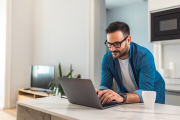 Focused man working over the laptop, leaning on a desk, with glasses on.