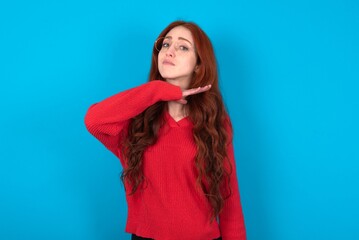 young woman wearing red sweater over blue background cutting throat with hand as knife, threaten...