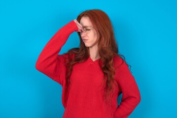 Sad young woman wearing red sweater over blue background suffering from headache holding hand on...