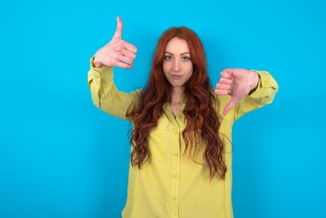 young woman wearing green sweater over blue background showing thumbs up and thumbs down, difficult...