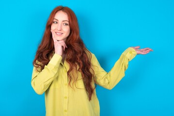 Funny young woman wearing green sweater over blue background holding open palm new product. I wanna buy it!