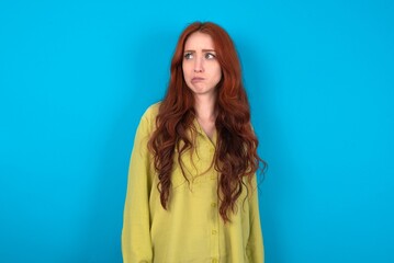 young woman wearing green sweater over blue background making grimace and crazy face, screaming out of control, funny lunatic expressing freedom and wild.