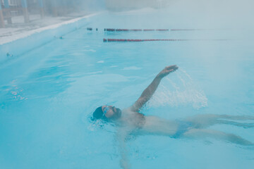 Fit swimmer male training swim backstroke in open winter swimming pool with fog. Geothermal outdoor...
