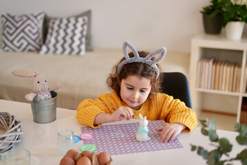 Cute little girl in bunny ears making decorations for Easter at home. Traditional Easter party preparation and decoration concept