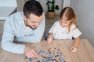 Smart game. Man and his daughter sitting at a table and looking at puzzle pieces while playing at home together. Focus on girl