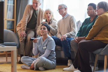 Diverse group of senior people watching TV in retirement home together with young nurse holding remote - obrazy, fototapety, plakaty