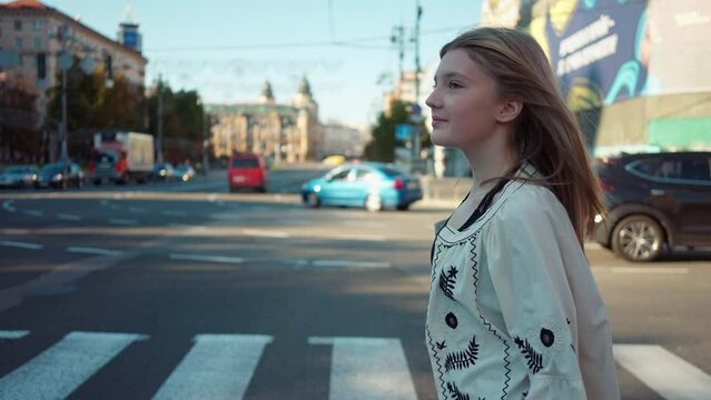 Side view tracking shot of smiling confident teenage girl crossing city street in slow motion. Live camera follows beautiful Caucasian teenager in embroidered shirt walking outdoors