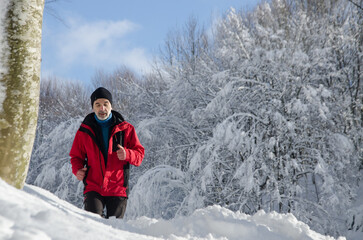 Fototapeta na wymiar Cross country running in winter. Happy fit senior man jogging in snowy forest on sunny morning. Sporty mature guy going for run in frosty weather, getting ready for race or marathon