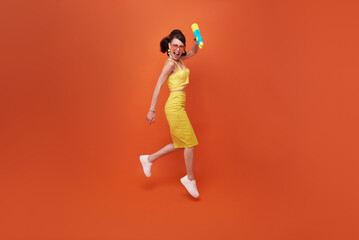 Fototapeta na wymiar Jumping tourist woman traveling with water gun during Songkran festival studio on copy space orange background.