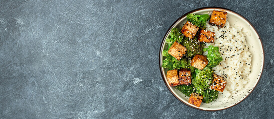 Asian vegan bowl with rice, broccoli and fried tofu on a dark background. Long banner format. top...