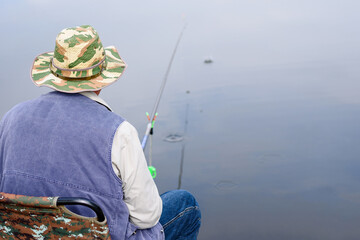 Elderly angler is fishing with fishing rod on lake sitting on chair. Leisure