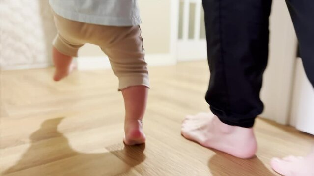 baby newborn first steps. mother teaches daughter baby to take the first steps with bare feet on the floor. baby walks barefoot on the floor at home light learning. baby newborn first steps indoors