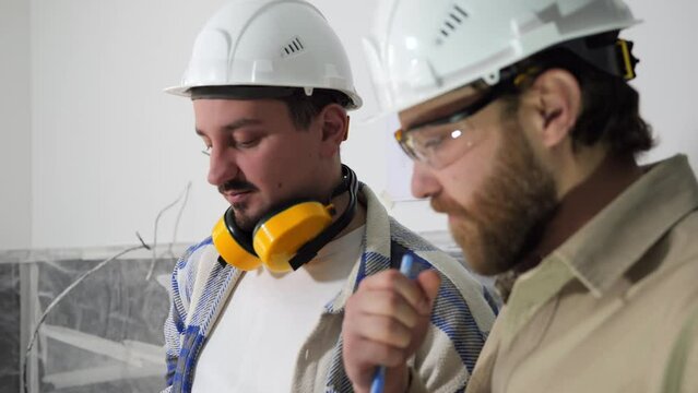 Two bearded construction fitters in white helmets are standing in the middle of the construction site, discussing construction documents and pointing to the interior of the apartment. High quality 4k