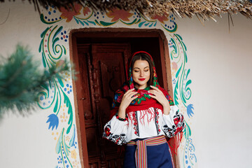 Charming smiling woman in traditional ukrainian handkerchief, necklace and embroidered dress standing at background of decorated hut. Ukraine, style, folk, ethnic culture
