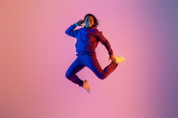 Fototapeta na wymiar Joyful energetic black woman in sportswear having fun and jumping high on neon colorful studio background, full length