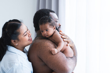 African Nigerian father holding cute newborn baby on shoulder in arms with Asian mother smiling looking at infant with love at home, standing near window. Multiracial family bonding and child care.