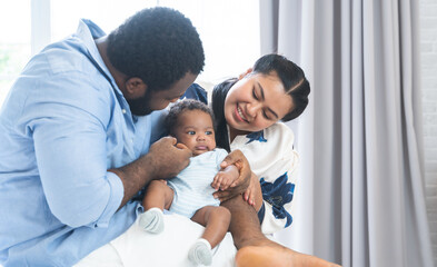 African father and Asian beautiful mother holding cute newborn baby in arms, playing and looking at infant with love and care at home. Multiracial family and child care concept