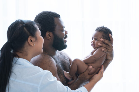 African Nigerian father and Asian mother carrying holding cute newborn baby in parents hands, smiling looking at infant with love at home, standing. Multiracial family bonding and child care concept