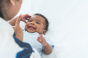 Happy African Nigerian newborn baby lying on white bed at home with mother talking and playing....