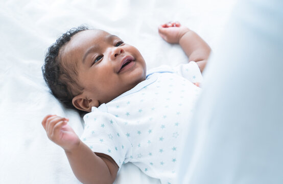 Happy African Nigerian newborn baby lying on white bed at home with mother talking and playing. Innocence infant with curly hair smiling at mom. Copy space