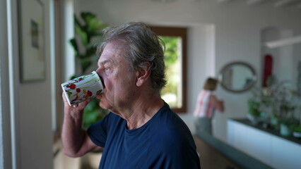 One mature older person drinks coffee or tea standing by window looking out in contemplation. 70s senior man with pensive expression