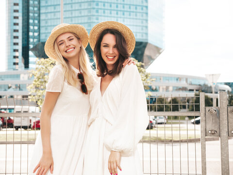 Two young beautiful smiling hipster female in trendy summer white dress clothes and straw hats. Sexy carefree women walking in street. Positive models having fun, hugging and laughing