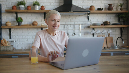 Healthy elderly woman in pink t-shirt using laptop, smiling and drinking orange juice