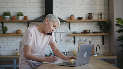 Elderly woman in pink t-shirt standing at the kitchen, talking on the phone and working on laptop