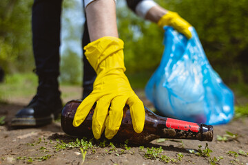 A young woman in yellow gloves collects abandoned garbage in a black bag in the forest. Plastic pollution and environmental protection