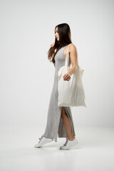 Brunette woman in grey dress holding cotton shopper bag with vegetables, products in white room....