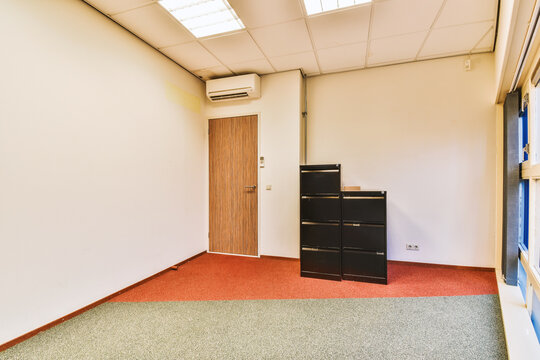 an empty office space with red carpet and white walls, there is a small black filing cabinet in the corner
