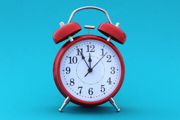 A red alarm clock stands on a blue background. The time shows five minutes to twelve	