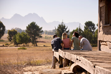 Rear View Of Group Of Friends On Vacation Sitting On Porch Of Countryside Cabin